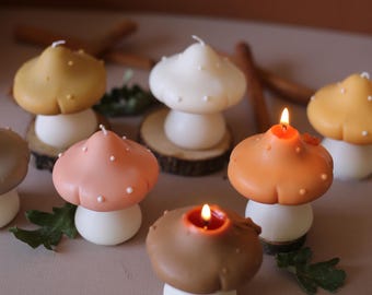 May include: A collection of mushroom-shaped candles in various colors, including brown, orange, pink, and yellow. Each candle has a white base and a textured cap with small white dots. Some candles are lit, with a warm glow.