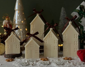 May include: Set of six white house-shaped candles with brown ribbon bows. The candles are arranged on a white surface with a snowy background.