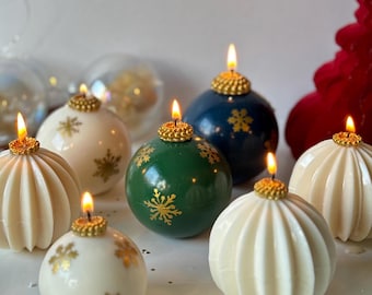 May include: Assortment of six white, green, and blue Christmas ornament candles with gold snowflake designs and a gold top. The candles are lit and arranged on a white surface.