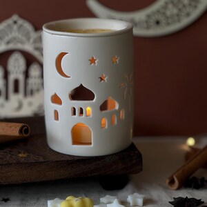 Peut inclure: Un photophore en céramique blanche avec des motifs de mosquée et d'étoiles découpés. Une lune et des étoiles sont découpées sur le côté. Une bougie allumée brille à l'intérieur, illuminant les détails. Le photophore est posé sur un plateau en bois.