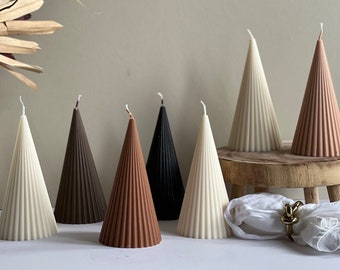 May include: Eight cone-shaped candles in various colours, including white, brown, black, and beige. The candles are arranged on a wooden surface and a white cloth.