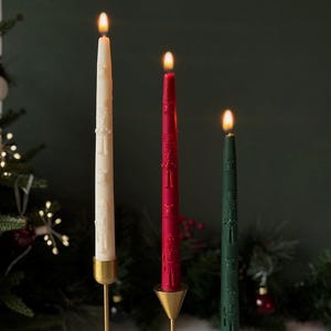 Pode incluir: Três velas cônicas acesas em creme, vermelho e verde, cada uma com um design em relevo detalhado. As velas estão em castiçais de metal dourado com base cônica. Decoração festiva de feriado.