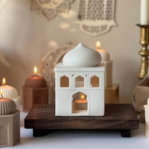 Peut inclure: Un photophore en céramique blanche en forme de mosquée, avec un dôme et des fenêtres en arc, contenant une bougie chauffe-plat allumée. Il repose sur un plateau en bois foncé, avec d'autres bougies en arrière-plan. Thème : décoration intérieure.