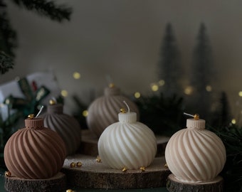 May include: Several decorative, swirled-design candles in shades of brown and white, shaped like Christmas ornaments. Each candle sits on a small wooden disc. Gold-colored beads are scattered around the candles, with a green background.