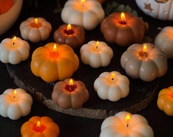 May include: A collection of pumpkin-shaped candles in various colors, including white, orange, and brown, with lit wicks. The candles are arranged on a dark surface, creating a warm, autumnal ambiance. Some candles are on a wooden slice, and a decorative pumpkin is in the background.