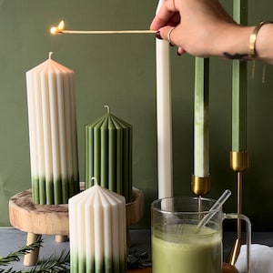 Pode incluir: Um conjunto de velas de pilar verdes e brancas com um design canelado. As velas est&atilde;o em um suporte de madeira e uma t&aacute;bua de cortar de madeira. H&aacute; um copo de ch&aacute; matcha verde na t&aacute;bua de cortar.