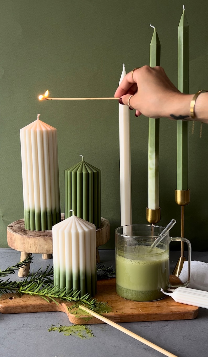 Pode incluir: Um conjunto de velas de pilar verdes e brancas com um design canelado. As velas est&atilde;o em um suporte de madeira e uma t&aacute;bua de cortar de madeira. H&aacute; um copo de ch&aacute; matcha verde na t&aacute;bua de cortar.
