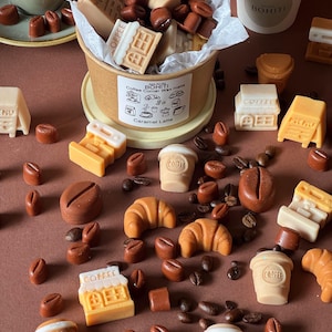 May include: A collection of wax melts shaped like coffee cups, croissants, and coffee shop storefronts, with the label "MAISON BOHITI Coffee Corner Wax melts Caramel Latte" on a paper cup, scattered with dark brown coffee bean-shaped wax pieces.