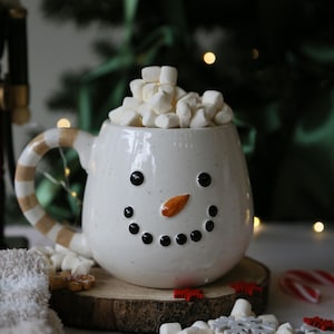 Peut inclure: Un mug en céramique blanche en forme de bonhomme de neige avec une finition mouchetée, des yeux et une bouche en points noirs, et un nez orange en forme de carotte. L'anse présente des rayures beiges et blanches. Rempli de mini guimauves.