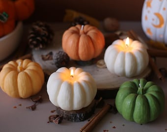 May include: A collection of pumpkin-shaped candles in various colors, including orange, white, and green. Some candles are lit, emitting a warm glow. The candles are arranged on a wooden surface with cinnamon sticks and decorative accents.