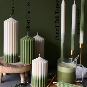 Pode incluir: Conjunto de velas caneladas verdes e brancas em v&aacute;rios tamanhos e estilos, incluindo velas caneladas grossas altas, velas caneladas grossas curtas, velas finas altas e velas caneladas finas. As velas s&atilde;o exibidas em uma superf&iacute;cie de madeira com uma x&iacute;cara de ch&aacute; verde.