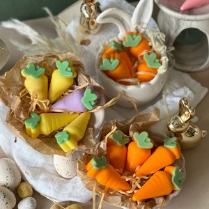 May include: A collection of handmade carrot-shaped soaps in various colours, including yellow, orange, and purple. The soaps are arranged in small baskets lined with brown paper and surrounded by speckled Easter eggs.