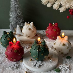 May include: A collection of spherical candles shaped like Christmas ornaments in red, green, and white. Each candle features a molded bow at the top and gold-colored snowflake and star accents. Some candles are lit, showing a small flame.
