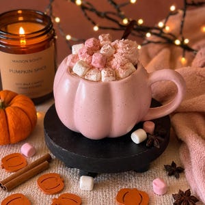 May include: A light pink ceramic mug shaped like a pumpkin, filled with a beverage topped with white and pink marshmallows and a dusting of cinnamon. The mug sits on a black circular stand surrounded by small orange wooden pumpkin cutouts.