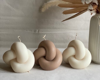 May include: Three knotted candles, one brown and two white, on a light grey surface. The candles are arranged in a row.