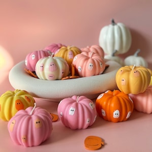 Könnte beinhalten: Eine Sammlung von kürbisförmigen Kerzen in verschiedenen Farben, darunter Rosa, Orange, Gelb und Weiß. Jede Kerze hat ein Geistergesicht-Design. Einige Kerzen sind in einer weißen Schale angeordnet, andere auf einer rosa Oberfläche. Einige kleine orangefarbene Kürbis-Ausschnitte sind ebenfalls vorhanden.