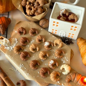 Könnte beinhalten: Nahaufnahme eines Holzschneidebretts mit einer Vielzahl kleiner, brauner, croissantförmiger Gebäckstücke. Die Gebäckstücke sind in einem Kreis um einen kleinen, weißen, hausförmigen Wachsschmelzer angeordnet. Der Wachsschmelzer hat eine rote Wachsschmelze im Inneren. Im Hintergrund befinden sich auch Zimtstangen und ein braunes Croissant.