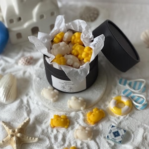 Puede incluir: Una caja cilíndrica negra llena de ceras aromáticas en forma de cangrejo, blancas y amarillas, forrada con papel de seda blanco. La etiqueta de la caja dice "BOHITI Crab Waxmelts". Varias ceras de cangrejo sueltas están esparcidas alrededor.