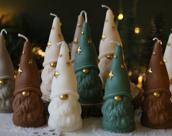 May include: A set of nine gnome-shaped candles in various colors, including brown, white, and green. Each candle has a pointed hat with gold stars and a gold nose. The candles are arranged on a white surface.