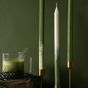 Pode incluir: Tr&ecirc;s candelabros altos com velas verdes e brancas sobre uma base dourada. Os candelabros est&atilde;o sobre uma mesa de madeira com um fundo verde.