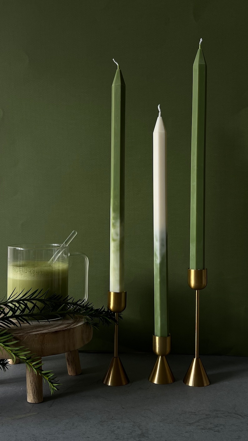 Pode incluir: Tr&ecirc;s candelabros altos com velas verdes e brancas sobre uma base dourada. Os candelabros est&atilde;o sobre uma mesa de madeira com um fundo verde.