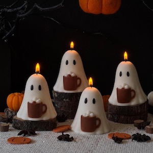 May include: Four white ghost-shaped candles, each featuring two black oval eyes and holding a brown mug filled with white marshmallows. Each candle is lit with a warm flame and sits atop a circular wooden log slice base.
