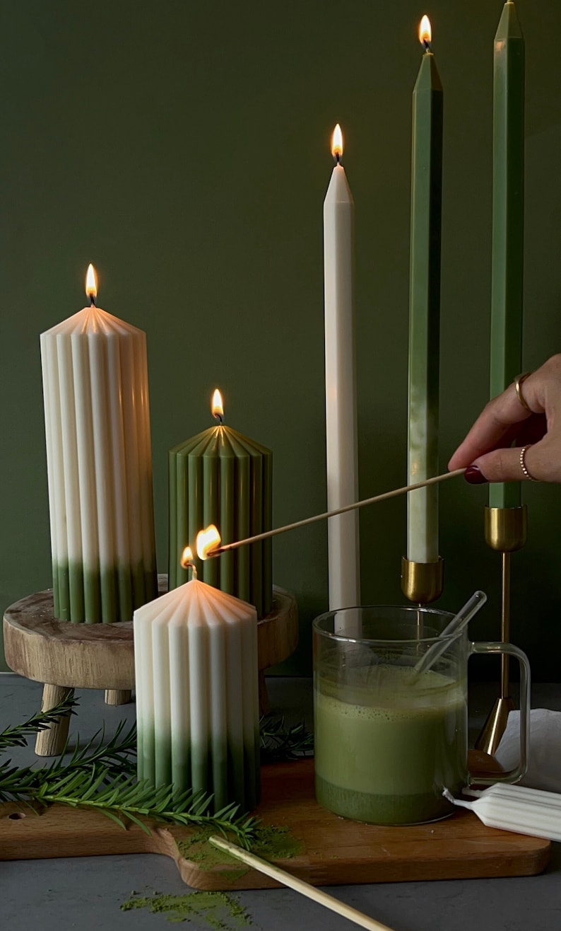 Pode incluir: Quatro velas, duas verdes e duas brancas, com um efeito degrad&ecirc; verde, est&atilde;o acesas e dispostas sobre uma superf&iacute;cie de madeira. Uma caneca de vidro cheia de uma bebida verde fica na frente das velas. P&oacute; verde &eacute; espalhado sobre a superf&iacute;cie.