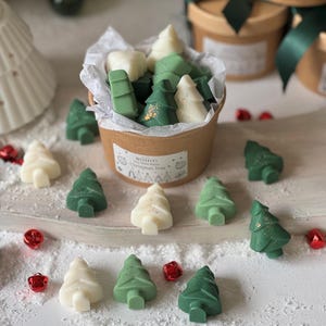 May include: A collection of Christmas tree-shaped wax melts in shades of green and white, some with gold accents, presented in a small brown container. Scattered around are additional tree-shaped melts and red jingle bells, creating a festive holiday scene.