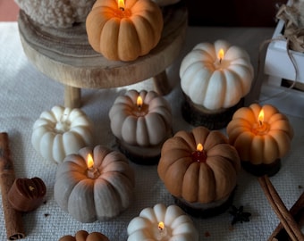 May include: Assortment of small pumpkin-shaped candles in various shades of brown, beige, and white. The candles are lit and have a warm glow. The candles are arranged on a white tablecloth with cinnamon sticks and star-shaped confetti.