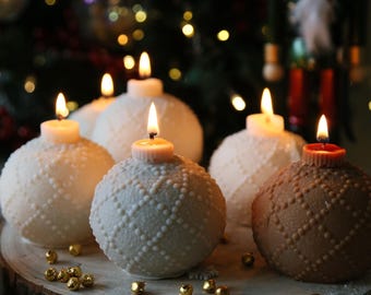 May include: Five white and one brown Christmas ornament candles with a textured surface and lit wicks. The candles are arranged on a wooden slice with gold beads scattered around them.