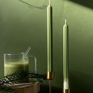 Pode incluir: Duas velas verdes com casti&ccedil;ais dourados sobre uma superf&iacute;cie cinzenta. As velas s&atilde;o altas e finas com uma textura canelada. Os casti&ccedil;ais s&atilde;o simples e elegantes com uma base c&oacute;nica.
