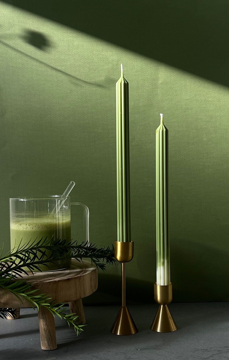 Pode incluir: Duas velas verdes com casti&ccedil;ais dourados sobre uma superf&iacute;cie cinzenta. As velas s&atilde;o altas e finas com uma textura canelada. Os casti&ccedil;ais s&atilde;o simples e elegantes com uma base c&oacute;nica.