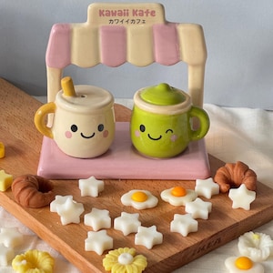 May include: A whimsical ceramic cafe scene with a pink and beige awning labeled "Kawaii Kafe." Two smiling mugs, one with a straw, sit beside decorative food-shaped wax melts, including stars, eggs, and a croissant, on a wooden board.