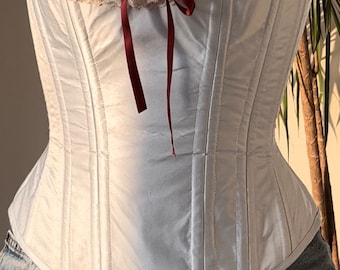 May include: A white satin corset with a lace and burgundy ribbon trim. The corset has a boned structure and a pointed bottom.