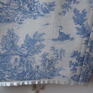 May include: A white fabric with a blue toile pattern featuring a cottage, trees, birds, and a heron. The fabric has a white satin trim.