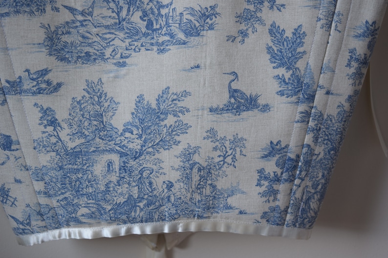 May include: A white fabric with a blue toile pattern featuring a cottage, trees, birds, and a heron. The fabric has a white satin trim.