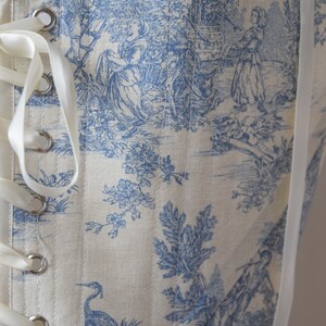 May include: A white corset with blue toile fabric featuring a pastoral scene with a river, trees, and figures. The corset has white satin ribbon lacing and metal eyelets.