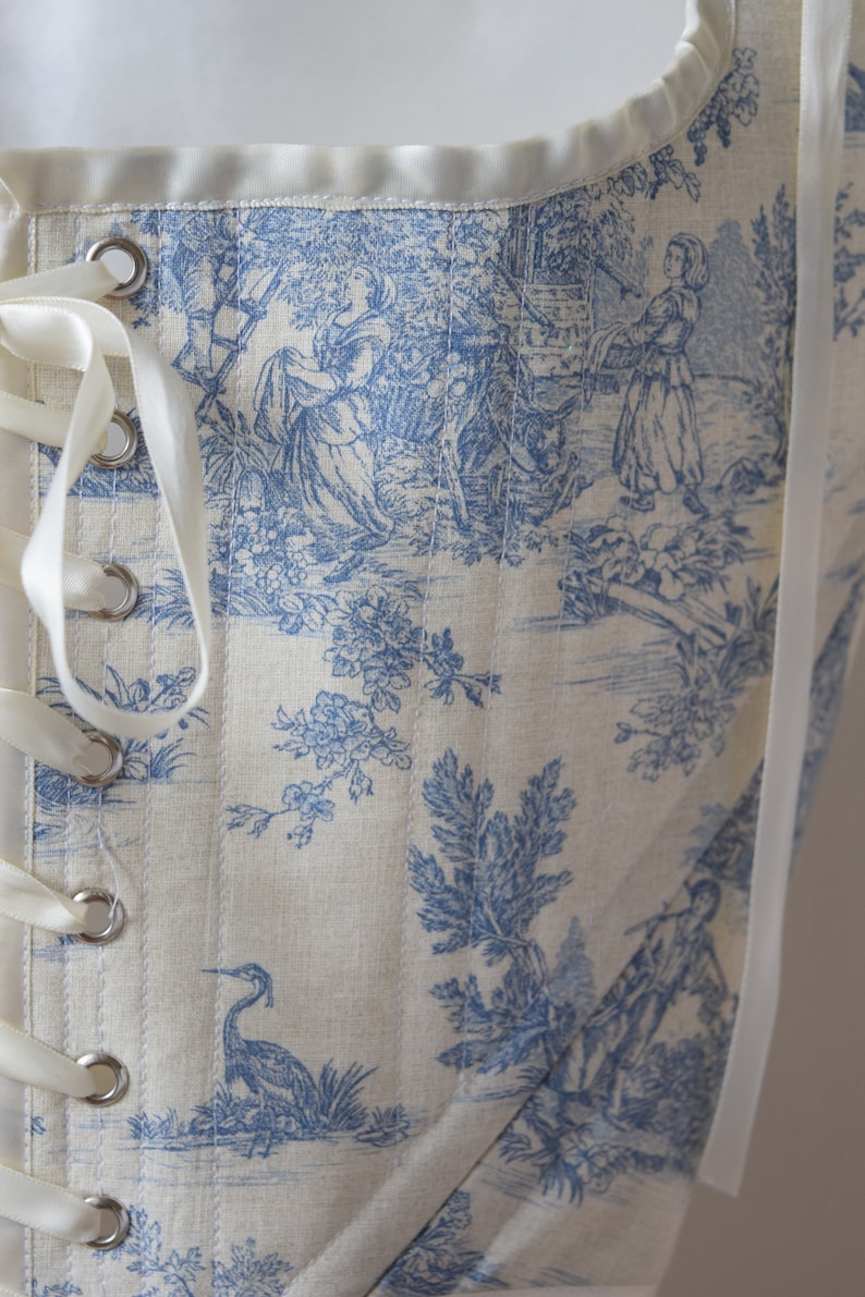 May include: A white corset with blue toile fabric featuring a pastoral scene with a river, trees, and figures. The corset has white satin ribbon lacing and metal eyelets.