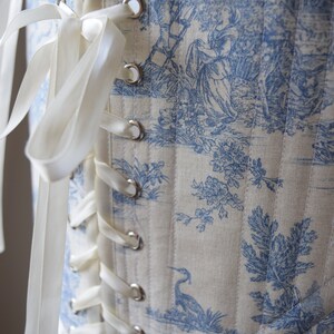 May include: A white corset with blue toile pattern and white satin ribbon lacing. The corset has a decorative trim at the bottom and silver eyelets.