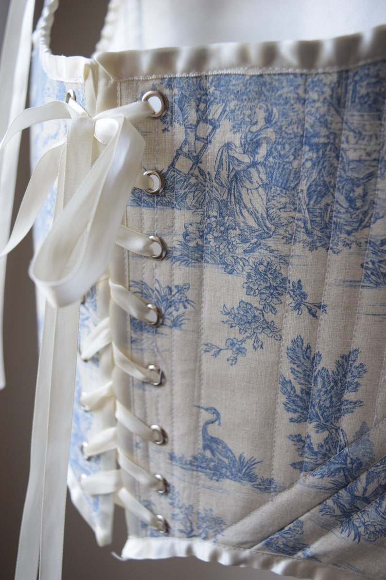 May include: A white corset with blue toile pattern and white satin ribbon lacing. The corset has a decorative trim at the bottom and silver eyelets.