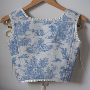 May include: A white sleeveless top with a blue toile pattern featuring a pastoral scene. The top has a white satin trim around the neckline and armholes.