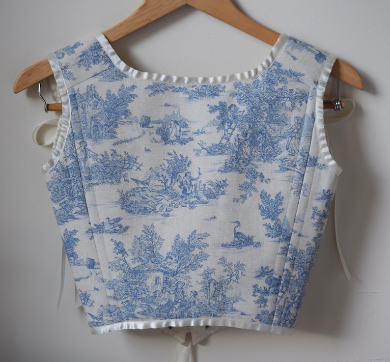 May include: A white sleeveless top with a blue toile pattern featuring a pastoral scene. The top has a white satin trim around the neckline and armholes.