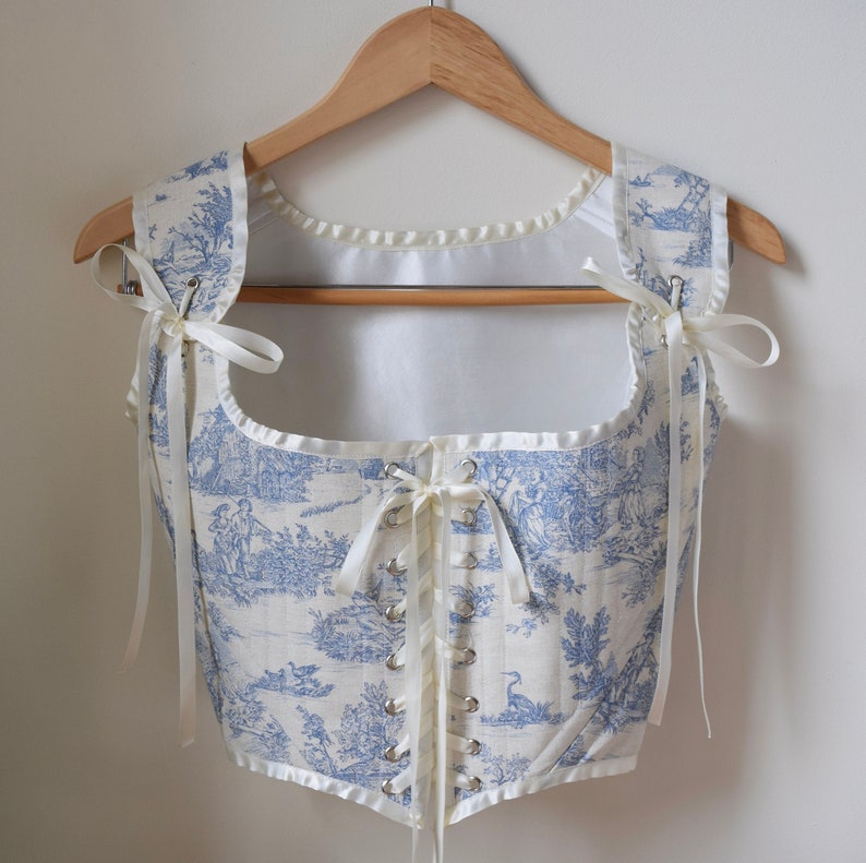May include: A white and blue toile patterned corset with a white satin trim and white ribbon ties. The corset has a lace-up back with silver eyelets.
