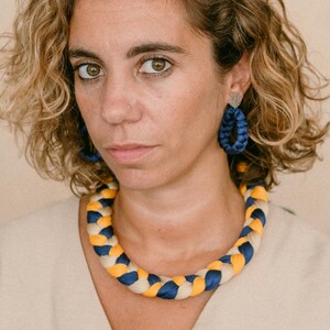 May include: A woman wearing a braided necklace and matching earrings. The necklace and earrings feature a pattern of blue, yellow, and cream-colored fabric. The woman is wearing a beige button-down shirt.