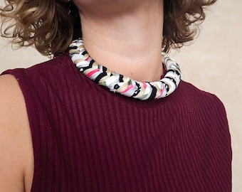 May include: A white, black and pink fabric necklace with a braided design. The necklace is worn by a person with curly hair and a maroon shirt.