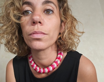 May include: A thick, braided statement necklace featuring a repeating pattern of satin-like ribbons in shades of bright pink, magenta, and cream. The necklace has a rounded, chunky silhouette that sits close to the neck, showcasing a woven texture.