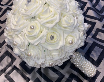 May include: A white bridal bouquet made of artificial roses with pearl and rhinestone accents. The bouquet has a pearl-covered handle.