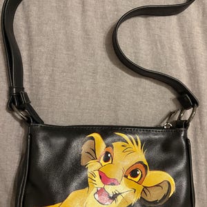 May include: Black shoulder bag with a colourful Simba illustration from The Lion King. The bag has a black strap and a zip. The Simba graphic is in yellow, orange, and brown.