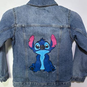 Peut inclure: Veste en jean bleu clair avec un dessin de personnage de dessin animé bleu et rose dans le dos. La veste a un col, une fermeture boutonnée et des manches longues. Le personnage est bleu avec de grandes oreilles.