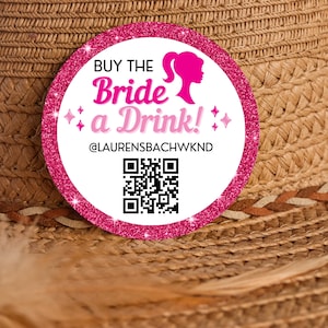 May include: Pink and white round sticker with the text "Buy the Bride a Drink!" and a QR code. The sticker is on a brown straw hat.